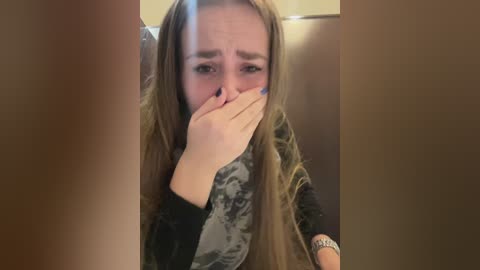 A video of a young woman with long, light brown hair, crying, covering her mouth with her right hand, wearing a camouflage-patterned scarf.