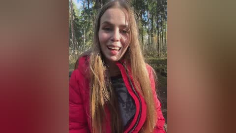 Video of a young Caucasian woman with long, straight, light brown hair, smiling. She wears a red puffy jacket over a dark shirt, standing in a forest with tall trees and green foliage.