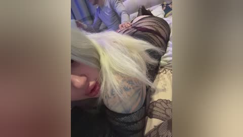 A video of a young woman with blonde hair, wearing a black fishnet top, lying on a bed. She has a tattoo on her arm and is partially visible. The background includes a blurry, softly lit room.