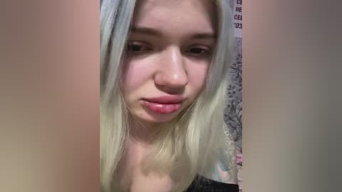 Video of a young woman with fair skin and long platinum blonde hair. She has a neutral expression, light pink lipstick, and a small nose ring. The background shows a patterned wall.