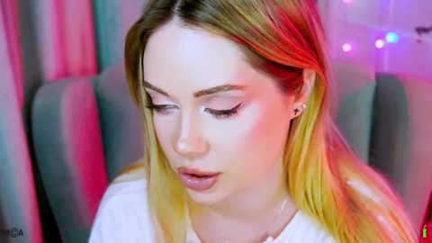 A video of a young woman with long, straight blonde hair, wearing makeup, including pink eyeshadow and mascara, sitting indoors with a colorful backdrop.