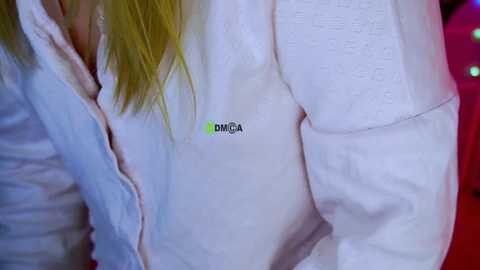 A video of a woman in a white bathrobe with a green logo on the left chest. Her blonde hair falls to the right. The background is blurred, featuring red and pink hues.