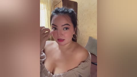 A video of a young Asian woman with dark hair, wearing a beige off-shoulder dress, adjusting her makeup in a dimly lit room with light-colored walls and a window.