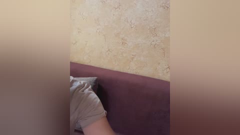 Video of a person lying on a purple sofa, head resting on a grey pillow, against a beige textured wallpaper wall.