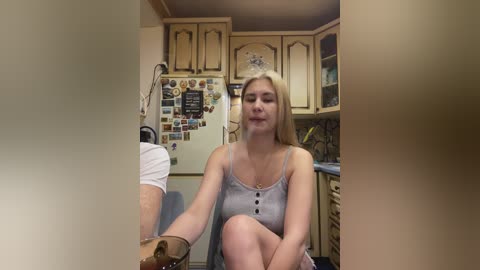 A young blonde woman in a gray tank top and shorts sits in a cozy kitchen, surrounded by wooden cabinets and a refrigerator adorned with magnets.