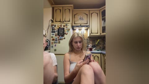 Video of a blonde woman in a strapless top, sitting in a cluttered, vintage-style kitchen with wooden cabinets and a refrigerator covered in magnets.