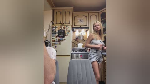 Video of a young blonde woman in gray shorts and a white tank top standing in a small, cluttered kitchen with beige cabinets, a fridge adorned with magnets, and a stove.
