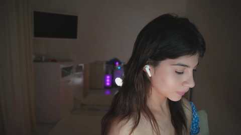 Video of a young woman with long dark hair, wearing white earbuds, standing indoors, looking downward. Background features a beige sofa, a TV, and a dimly lit room.