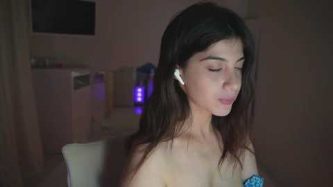 A video of a young woman with long, dark hair, wearing white earphones, seated in a dimly lit room with muted colors and a fridge in the background.