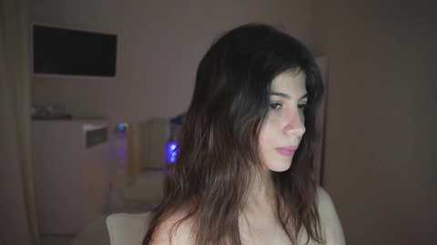 Video of a young woman with long, dark brown hair, fair skin, and light makeup, sitting indoors with a dimly lit background featuring a TV, a white wall, and a blue light.