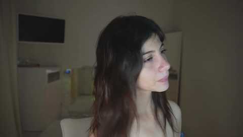 A video of a young woman with long, wavy dark hair, fair skin, and a slender build, seen in profile in a dimly lit room.