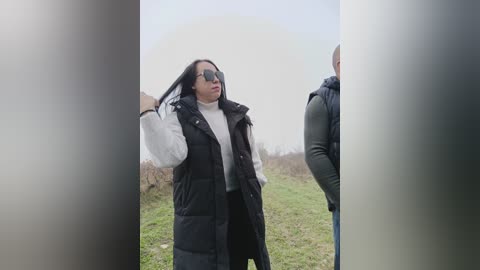 Video of a woman with dark hair and sunglasses, wearing a white turtleneck under a black puffer vest, standing in a grassy field with a man in a black hoodie, partially obscured by vertical blurs.