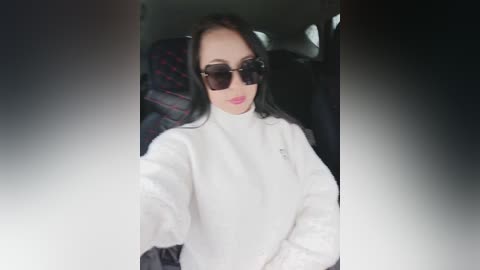 Video of a woman with long black hair, wearing oversized dark sunglasses, a white fluffy jacket, and sitting in a car with dark upholstery and a patterned seat cover.