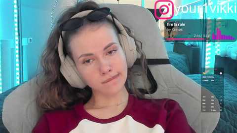 Video of a young woman with fair skin, brown hair, wearing a red shirt, sitting in a gaming chair, headphones on, in a modern, brightly lit room.