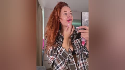 A video of a young woman with long, red hair, wearing a plaid shirt, applying lipstick in a mirror, with blurred walls and a closet in the background.