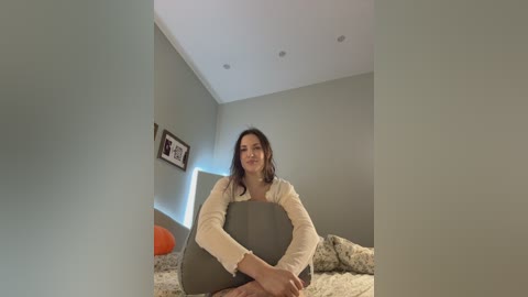 A video of a woman with dark hair, wearing a beige cardigan, sitting on a bed with a beige blanket. The room has light grey walls, a framed photo, and a soft orange ball.