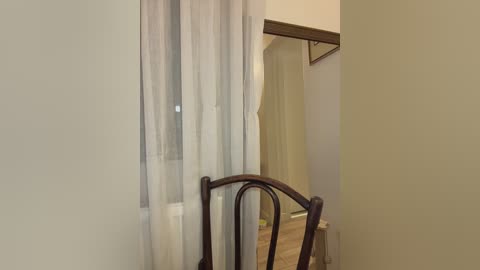 Video of a small, dimly lit room with a wooden chair, beige walls, and a window with sheer white curtains. The background shows an open doorway leading to another room.