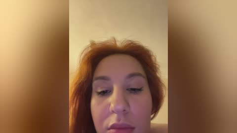 A close-up video of a light-skinned woman with long, reddish-brown hair, wearing minimal makeup, focusing on her face. The background is blurred and beige, emphasizing her eyes and lips.