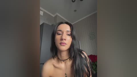 Video of a young woman with long, straight black hair and fair skin, wearing a black choker necklace, looking down, in a modern, neutral-toned room with a white ceiling and textured wallpaper.
