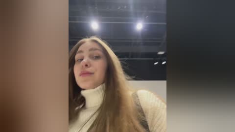 Video of a fair-skinned, blonde woman with long hair, wearing a cream-colored turtleneck sweater, in a dimly lit room with two bright overhead lights.