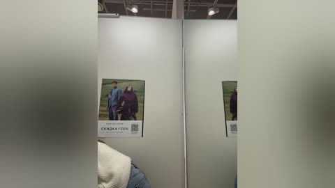 A video of a dimly lit, minimalist dressing room with two mirrors, showcasing a woman wearing a blue sweater, and a poster of a couple in outdoor clothing. The room has a white, industrial ceiling.