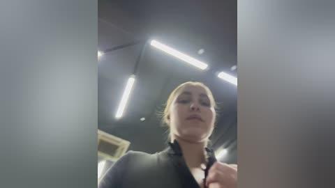 Video of a young woman with light skin, blonde hair tied back, wearing a black jacket, standing in a dimly lit room with fluorescent lights. Background shows part of a ceiling and wall.