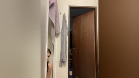 Video of a young man with short hair, wearing a grey bathrobe, standing in a white-tiled bathroom, with a wooden door and towel rack visible in the background.