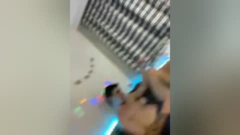 A blurry video showing a person, possibly a woman, wearing a black and white striped shirt, holding a phone with a colorful background, against a black and white checkered wall.