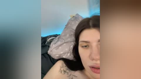 Video of a young woman with fair skin, dark hair, and light makeup, lying on a bed with a gray pillow and dark bedding, partially obscured by a blurred, brown-skinned person.