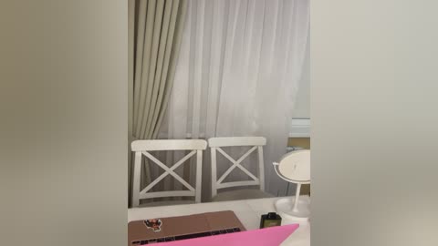 A video of a minimalist bedroom with white walls, a white bed, and grey curtains. A white chair with a cross design stands in the background. The room's light and neutral color scheme creates a calm atmosphere.