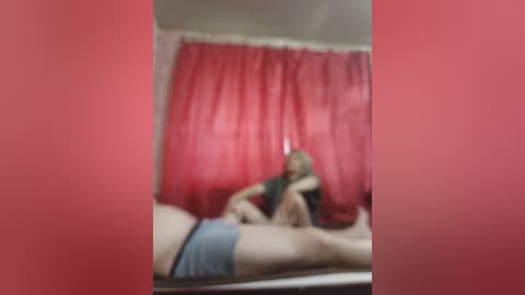 A blurred video of a person in a pink room, lying on a bed with a red curtain background, wearing blue shorts and a black top.