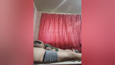 A video of two people lying on a bed in a dimly lit room with red curtains. The person on the left has short blonde hair, wears a green shirt, and gray shorts. The person on the right, with dark hair, is wearing a green shirt.