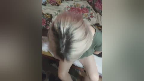 Video of a person with blonde hair in a messy bun, wearing a green shirt, standing in a room with a floral-patterned bedspread in the background.