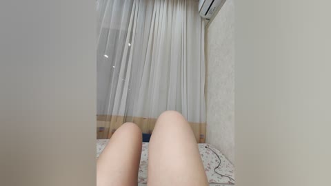 A video of a person's legs, pale skin, lying on a bed with white curtains and a floral-patterned sheet in a simple, well-lit room.