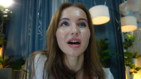 A video of a Caucasian woman with long brown hair, fair skin, and blue eyes, wearing a white lace top, smiling widely. Background features blue curtains, green potted plants, and modern light fixtures.