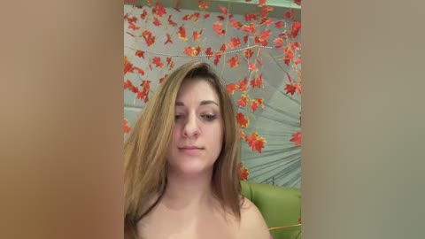Video of a topless young Caucasian woman with light skin and long brown hair, posing indoors against a backdrop of orange leaf garlands and a green couch.