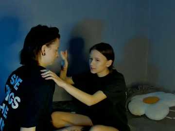 Video of two women in a dimly lit room with blue-gray walls. One woman, with short hair and wearing a black t-shirt, is sitting and touching the other's face. The other woman, with long hair and a black top, sits on the floor. A white flower decorates the background.