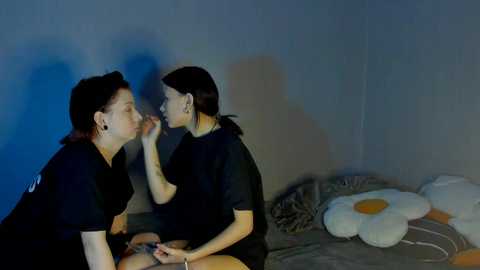 Video of two young women in black outfits, one applying lipstick to the other's lips, in a dimly lit room with a floral-patterned bedspread.