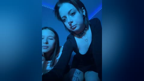 Video of two young women with pale skin and light brown hair, wearing black tops, posing against a blue-lit wall, both with closed eyes, creating a dreamy, ethereal atmosphere.