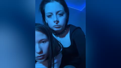 Video of three young women with pale skin and dark hair, wearing black clothing, under blue lighting, giving serious expressions, with one woman in the foreground.