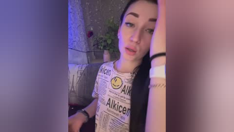 A video of a young woman with long black hair, wearing a New York Times T-shirt, sitting in a dimly lit room with a plant in the background.