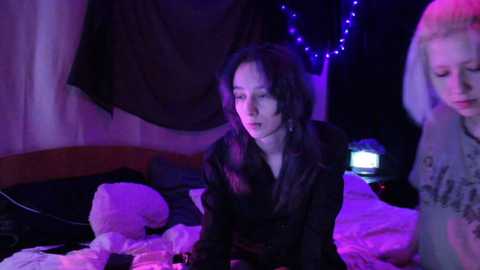 Video of three women in a dimly-lit bedroom with purple lighting. One woman with dark hair and a hoodie sits on a bed, another with light hair stands in the background, and a third woman with light hair is partially visible.