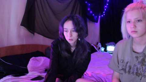 Video of two young women in a dimly lit bedroom; one with long black hair, wearing a black sweater, the other with short platinum hair, wearing a gray graphic tee.
