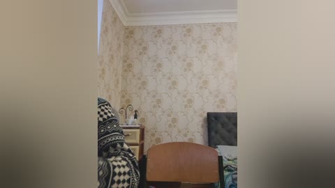 Video of a cozy bedroom with beige floral wallpaper, a tufted brown headboard, a black-and-white patterned blanket, and a dresser holding a lamp and clock.