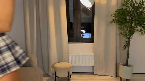 Video of a modern, minimalist living room featuring beige curtains, a white radiator, a small beige stool, a leafy plant in a white pot, and a person in a blue and white plaid shirt.