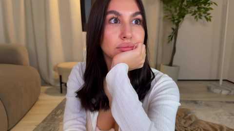 Video of a young woman with long, straight dark hair, light skin, and blue eyes, wearing a white cardigan, resting her chin on her hand, looking thoughtful in a modern living room with beige furniture and a potted plant.
