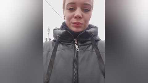 Video of a young woman with short brown hair, wearing a black puffy jacket with a high collar, standing in a dimly lit corridor. Her face is neutral, and the background is blurred.