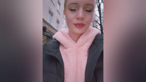 Video of a young woman with blonde hair, wearing a pink scarf, black jacket, and red lipstick, smiling, outdoors against a building.