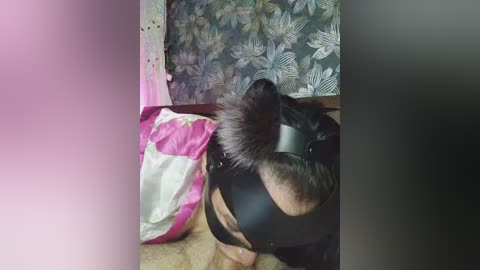 Video of a person in a black leather hood with a fluffy, black dog in a pink and white blanket, against a floral-patterned wall.