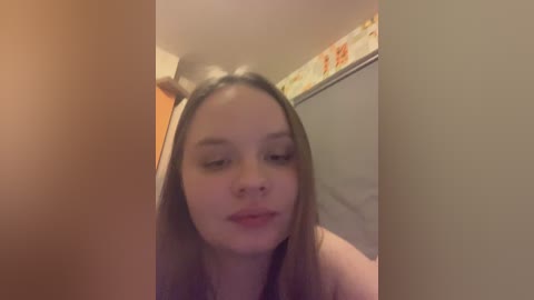 A video of a young woman with fair skin and long, straight brown hair, wearing a black top, captured indoors. She has a relaxed expression. The background features a beige wall with floral wallpaper and a shower curtain.